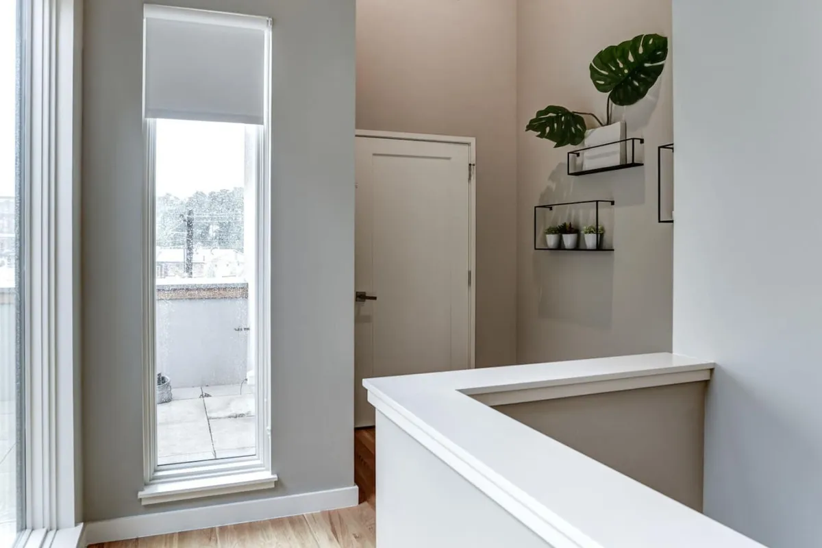A bright, airy interior space with a large window letting in abundant natural light, a green plant set on a shelf, and a clean white counter. The view through the window subtly suggests the presence of the lively cityscape outside.