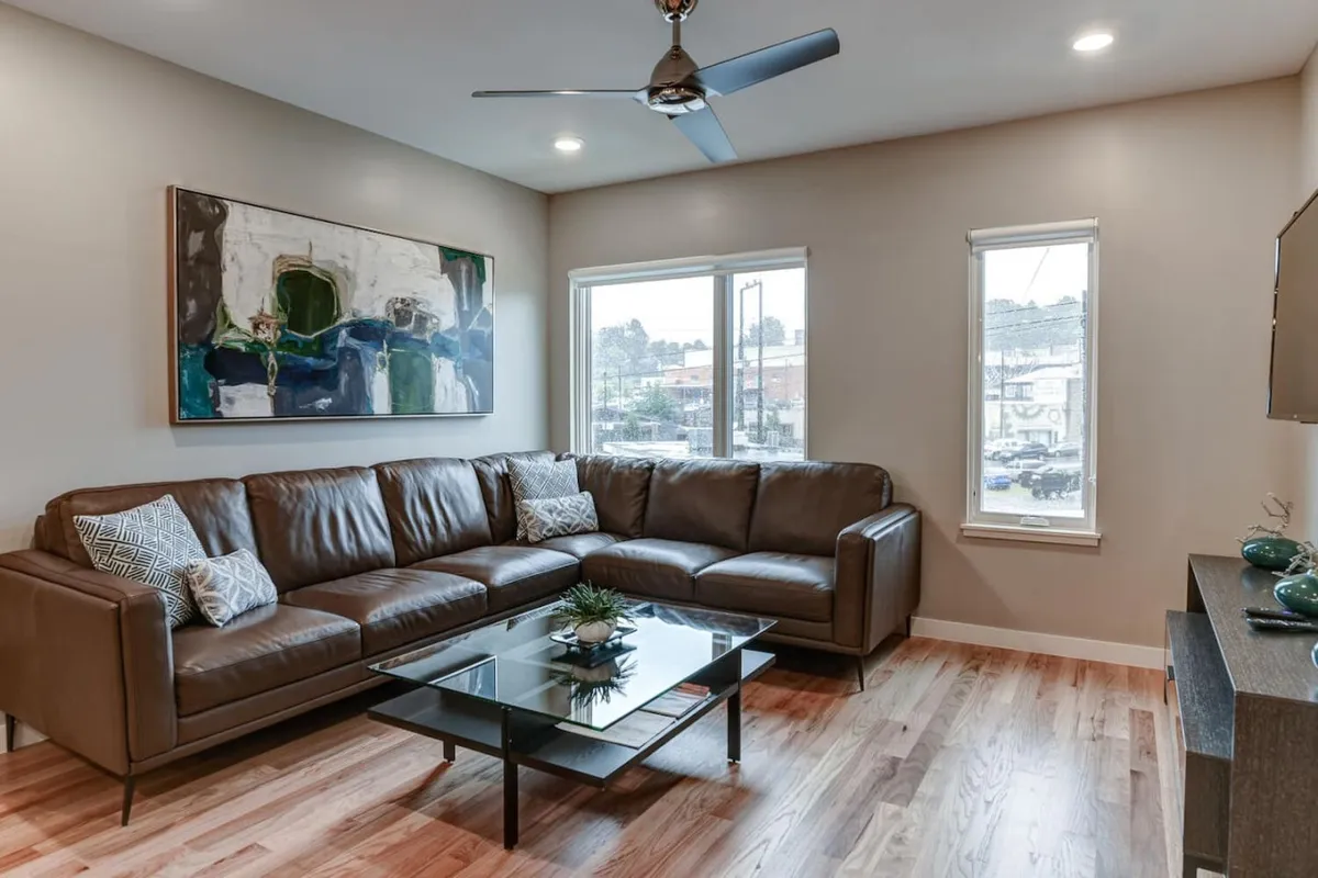 A spacious living area featuring a leather sectional sofa, glass coffee table, colorful abstract artwork, and expansive windows that fill the room with natural light. The arrangement invites groups to gather for conversation or a fun evening together.