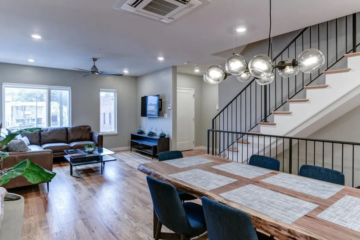 This image captures an open living and dining space featuring a cozy sofa, a contemporary dining table, a striking staircase, statement lighting, and a TV. Natural light fills the room, highlighting the inviting, modern atmosphere ideal for socializing and unwinding.