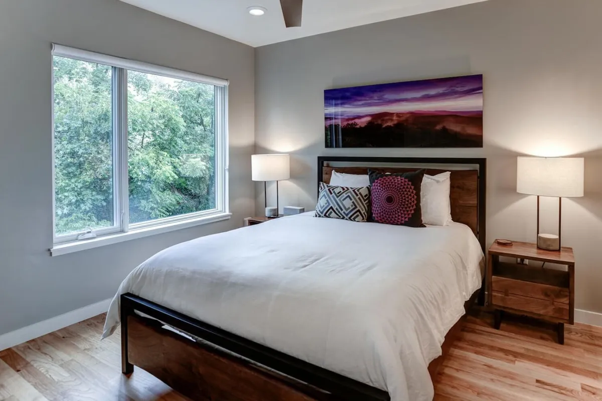 A serene bedroom with a cozy bed, window allowing in soft daylight, elegant artwork, and well-placed nightstands with lamps. The decor feels inviting and restful.