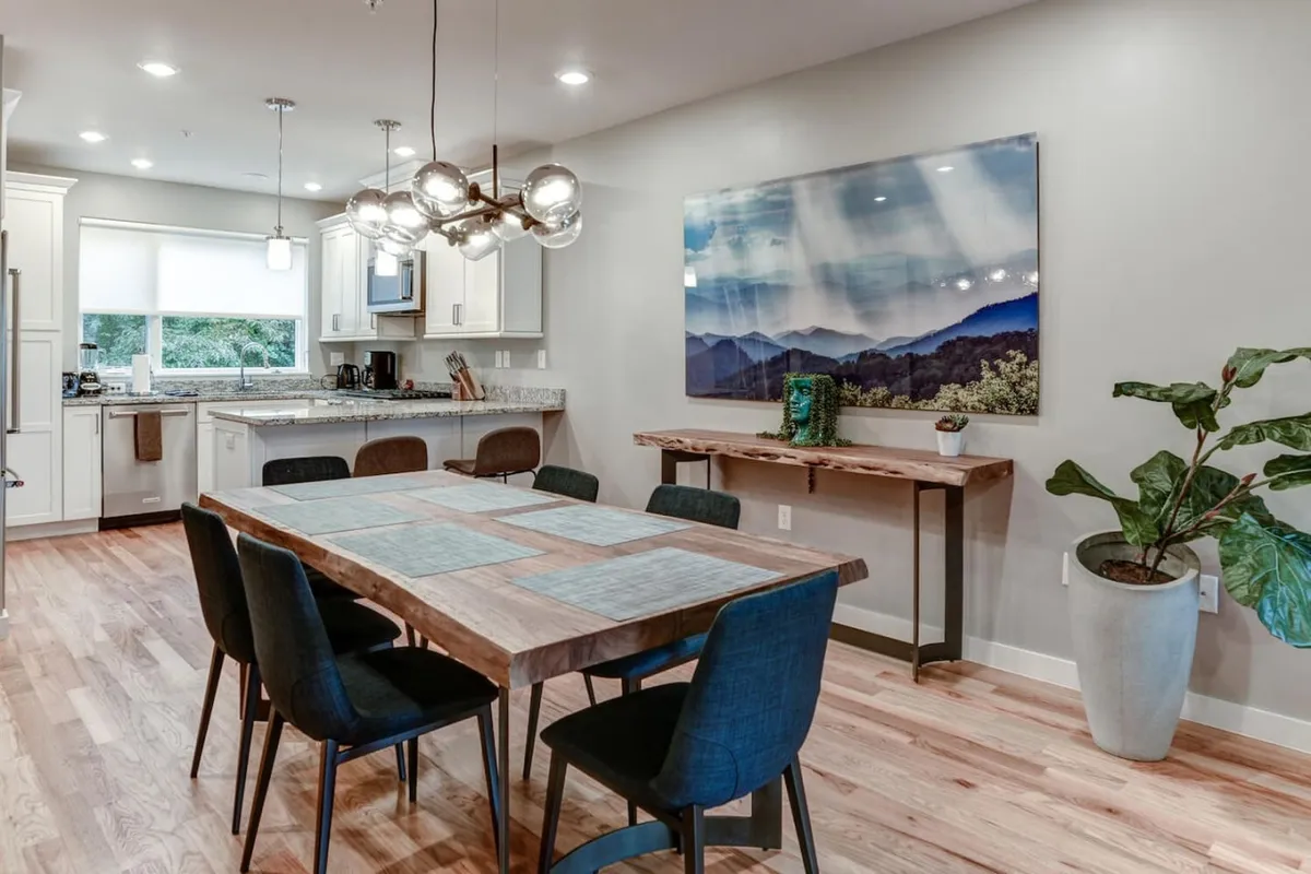 A contemporary dining area with a large table, adjacent kitchen island, bold artwork, and healthy houseplant. The setup invites shared meals, celebrations, and casual gatherings in a bright, open setting.