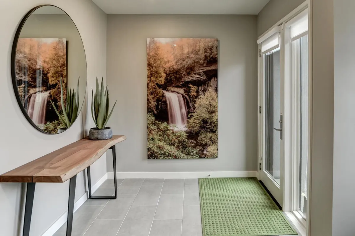 A welcoming entryway with a large circular mirror, wooden console table, waterfall artwork, a potted plant, and a green mat. The space exudes a calm, inviting atmosphere, setting the tone for the rest of the home.