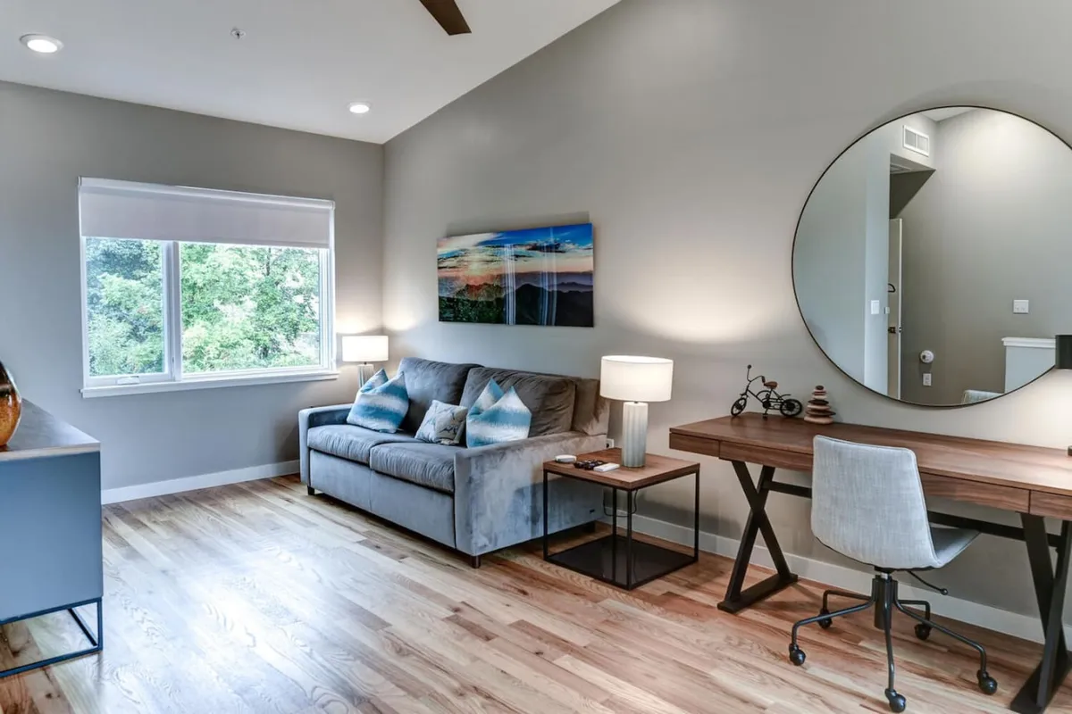 A bright and modern living area featuring a comfortable sofa, a stylish desk and chair setup, inviting artwork on the wall, a lamp, and a large window that lets in natural light and offers a view outside. The scene blends elements conducive to both productivity and relaxation.