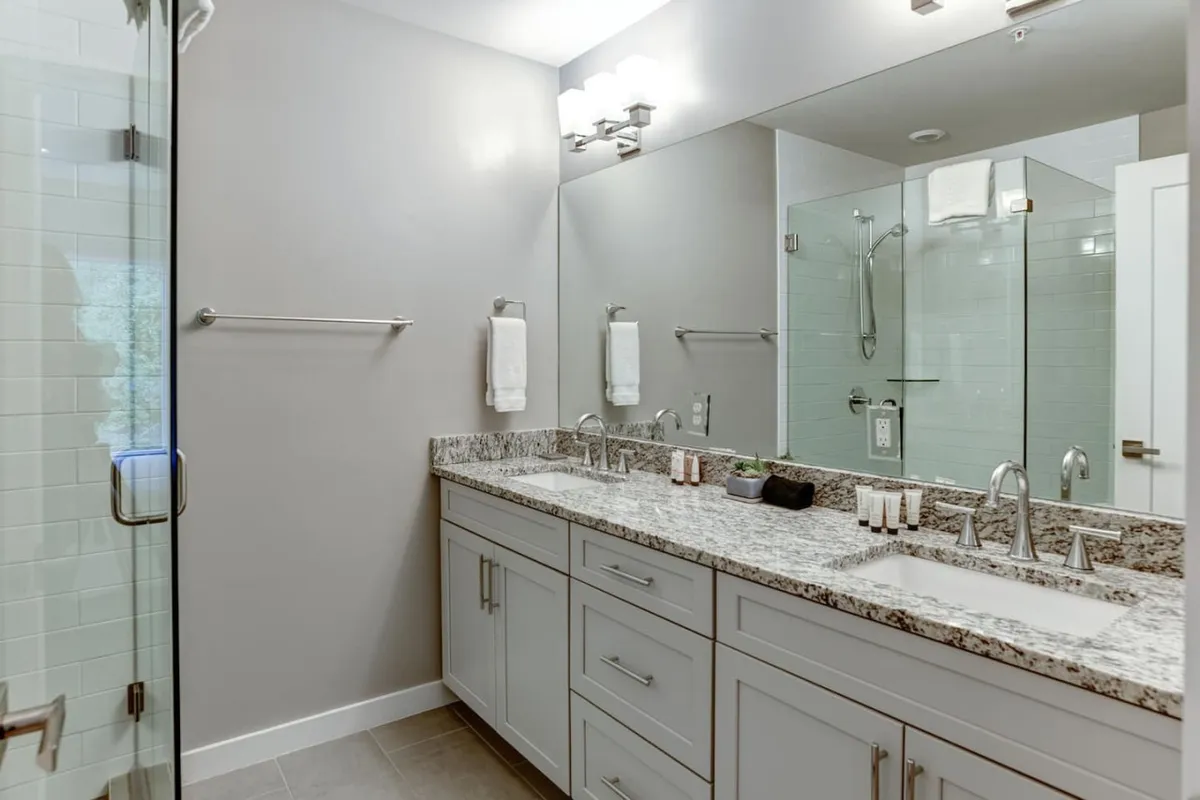 A bathroom with a double sink granite countertop, large mirror, and glass shower enclosure. The atmosphere is serene and spa-like, suitable for relaxing morning or evening routines.
