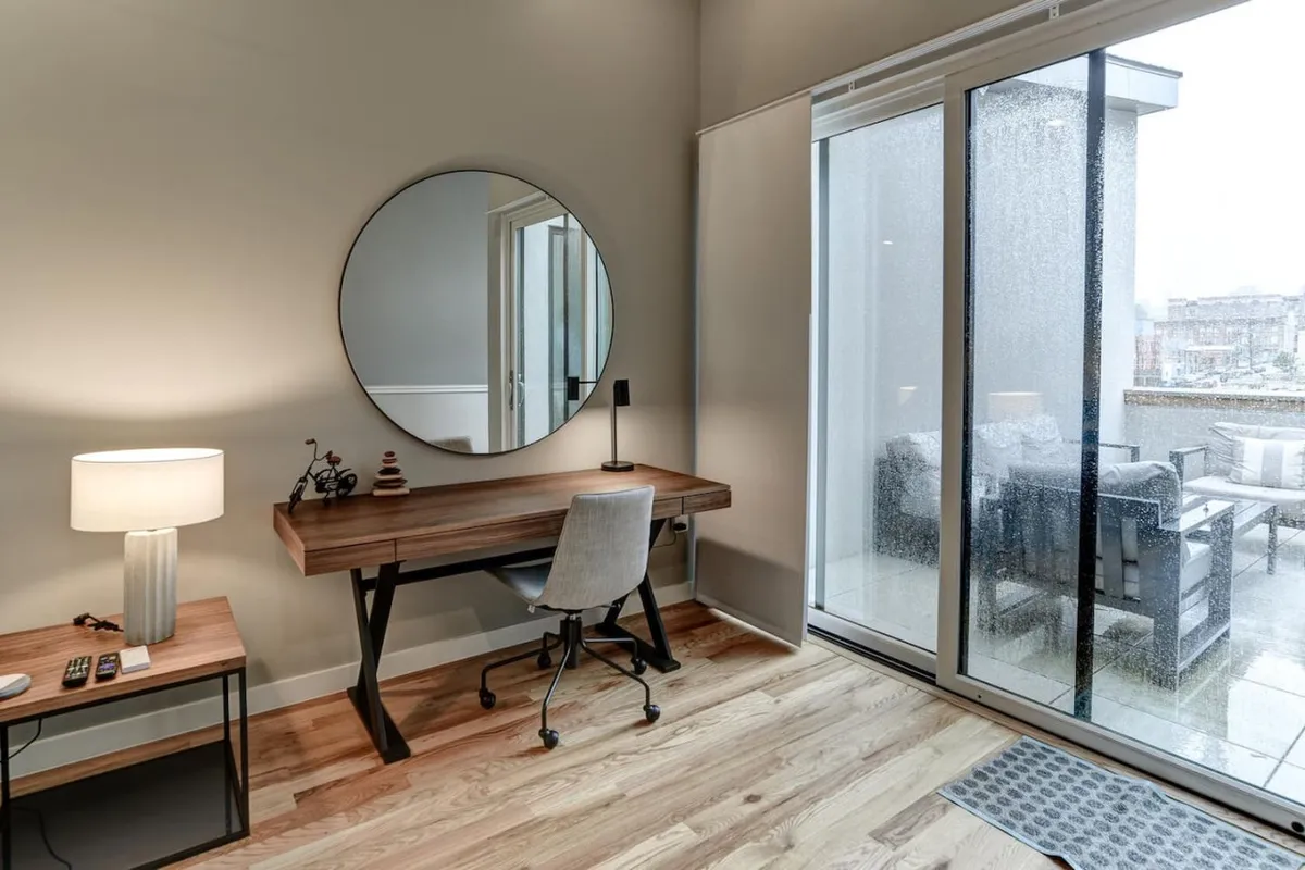 A versatile loft space featuring a desk for focused work, a mirror, a sliding glass door for natural light, and a lamp. The setting feels both functional and relaxing, ideal for a flexible sleeping or workspace.