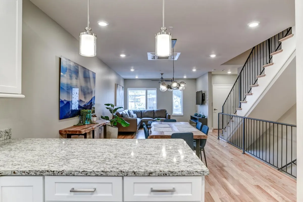 A perspective into a cohesive living, dining, and kitchen space, featuring a granite countertop, modern lighting, curated artwork, a comfortable sofa arrangement, and a welcoming dining table. The setting blends elegance with comfort, perfect for entertaining.
