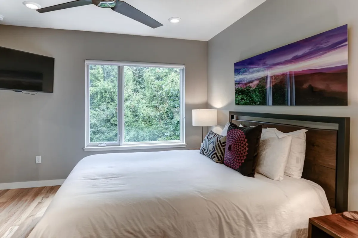 A tranquil bedroom featuring a bed adorned with decorative pillows, large window overlooking lush greenery, and tasteful wall art. The room exudes a peaceful ambiance with natural light streaming in.
