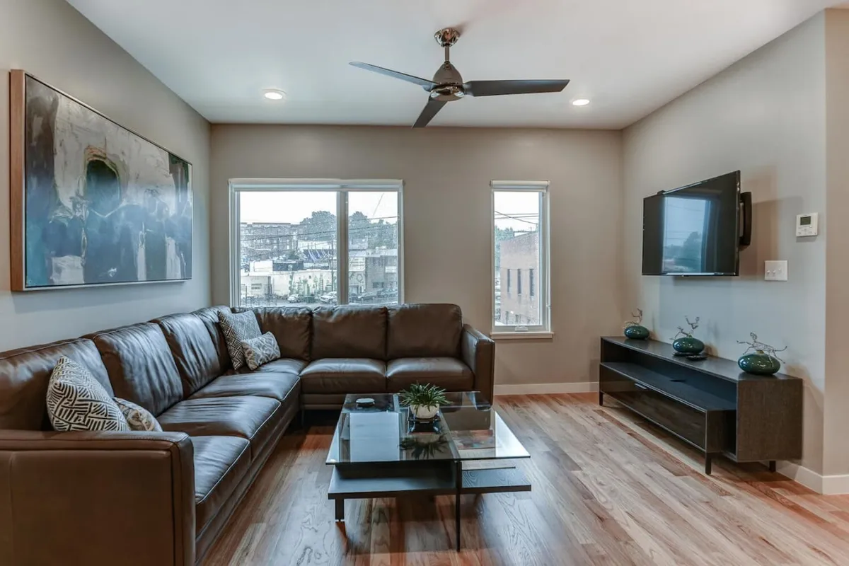 This image showcases a spacious living area with a leather sectional sofa, glass coffee table, large wall art, and a television mounted on the wall. The space is bathed in natural light and arranged for both relaxation and social gatherings.